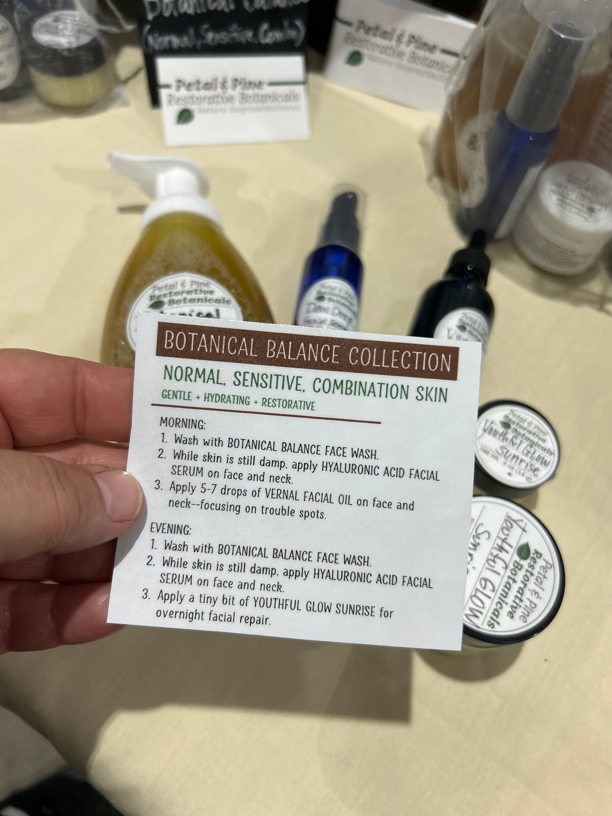 A hand holds a white card detailing the 'Botanical Balance Collection' face wash instructions, with various skincare bottles and jars blurred in the background on a beige countertop.