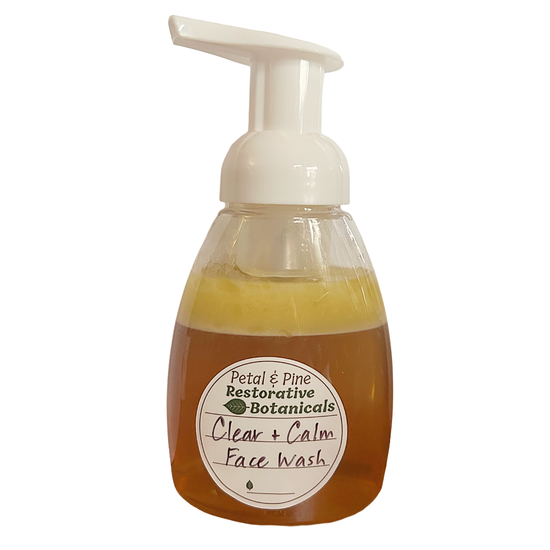 A clear plastic pump bottle of Petal & Pine Restorative Botanicals Clear + Calm Face Wash, filled with amber-colored liquid and topped with a white foam pump dispenser, shown against a plain white background.