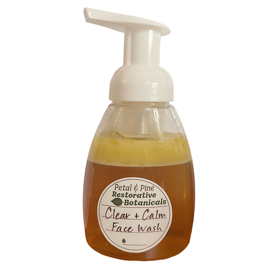 A clear plastic pump bottle of Petal & Pine Restorative Botanicals Clear + Calm Face Wash, filled with amber-colored liquid and topped with a white foam pump dispenser, shown against a plain white background.