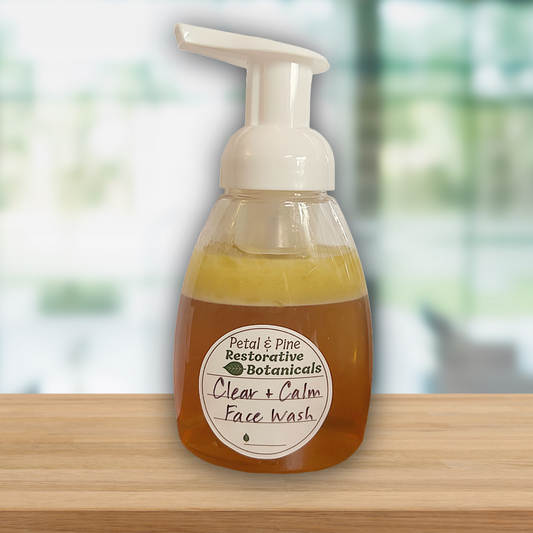 A clear plastic pump bottle of face wash with a white pump dispenser, sitting on a light wood surface against a softly blurred indoor background. The liquid inside is amber-brown, and the front label reads 'Petal & Pine Restorative Botanicals Clear + Calm Face Wash.