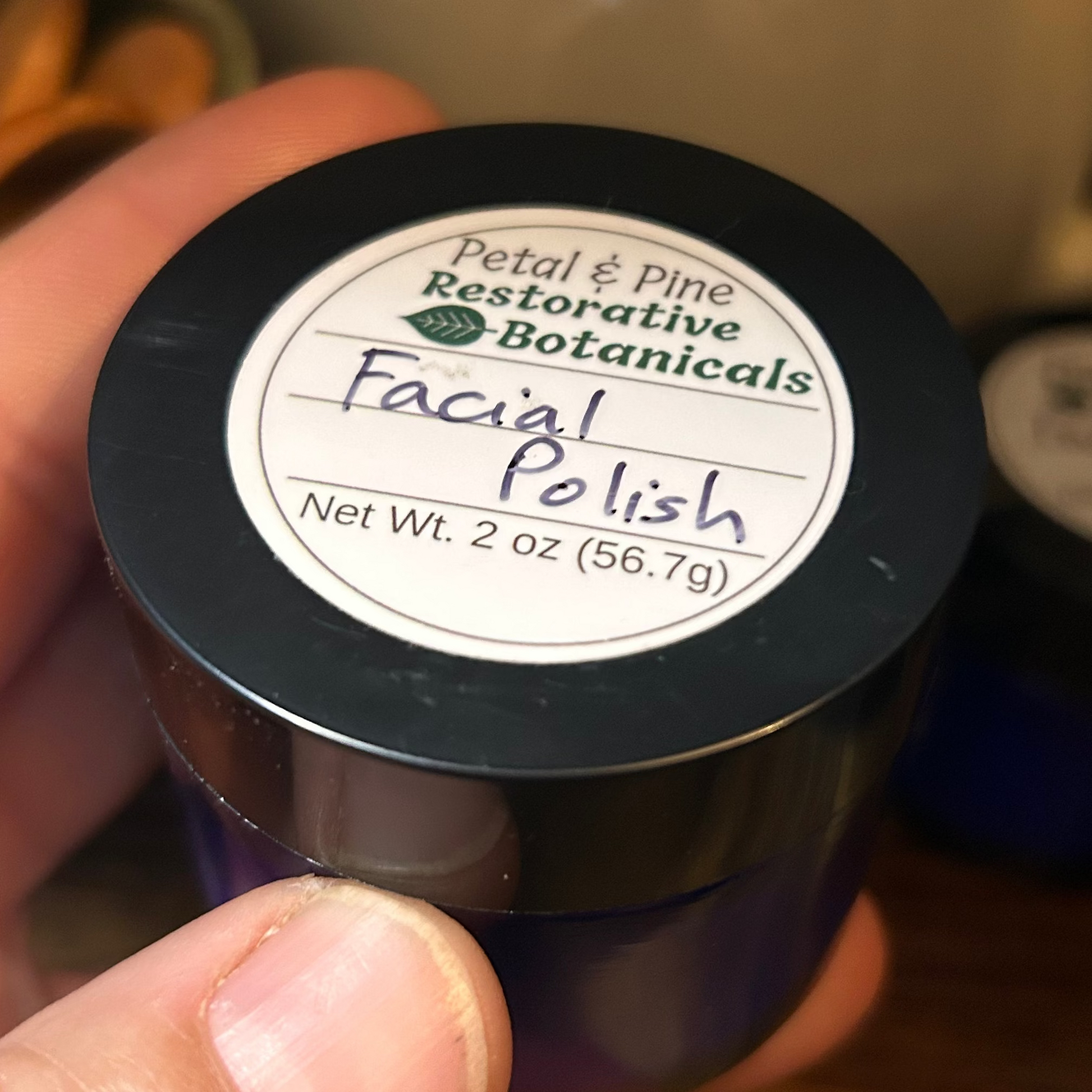 A hand holding a small dark jar of Petal & Pine Restorative Botanicals Facial Polish on a wooden table with a ceramic pitcher in the background.