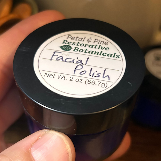 A hand holding a small dark jar of Petal & Pine Restorative Botanicals Facial Polish on a wooden table with a ceramic pitcher in the background.