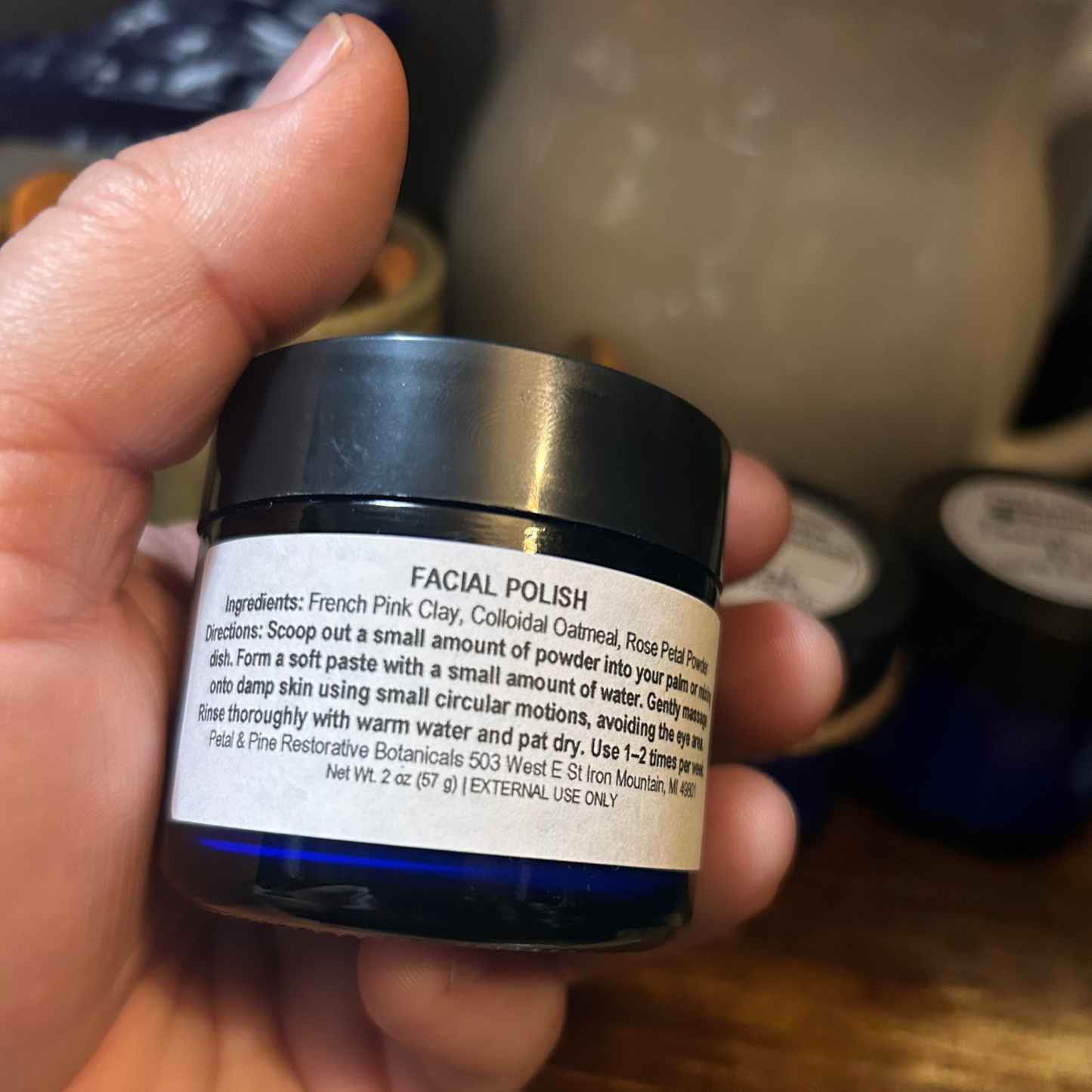 A hand holding a small black jar of facial polish with a white label, photographed over a wooden surface with other jars blurred in the background.