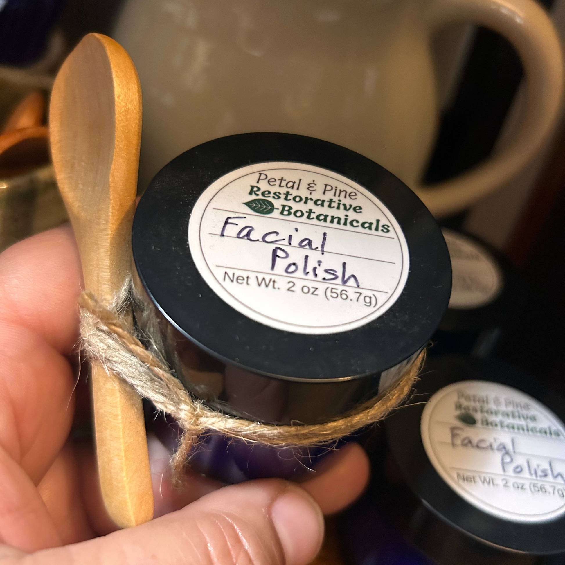 A hand holds a small jar of Petal & Pine Restorative Botanicals Facial Polish wrapped in twine, with a wooden spoon resting on top. The jar has a black lid and a white label, with another jar in the background and a ceramic mug behind it.