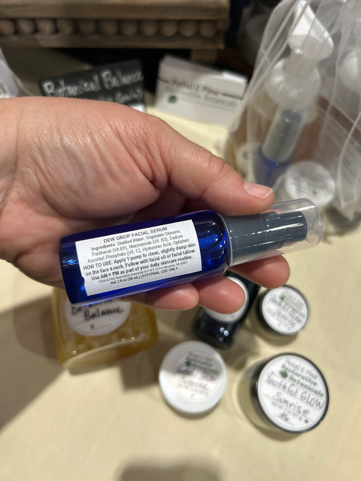 A hand holding a small blue bottle with a clear cap and a white label, with other skincare products blurred in the background on a beige tabletop.