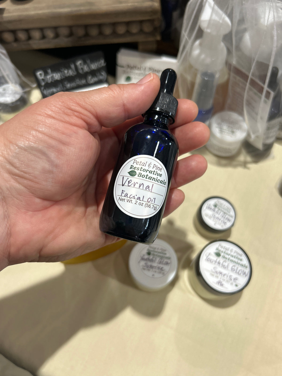 A hand holding a small dark cobalt glass dropper bottle labeled 'Vernal Fagaceae' by Botanical Balance, with other skincare products blurred in the background on a beige tabletop.