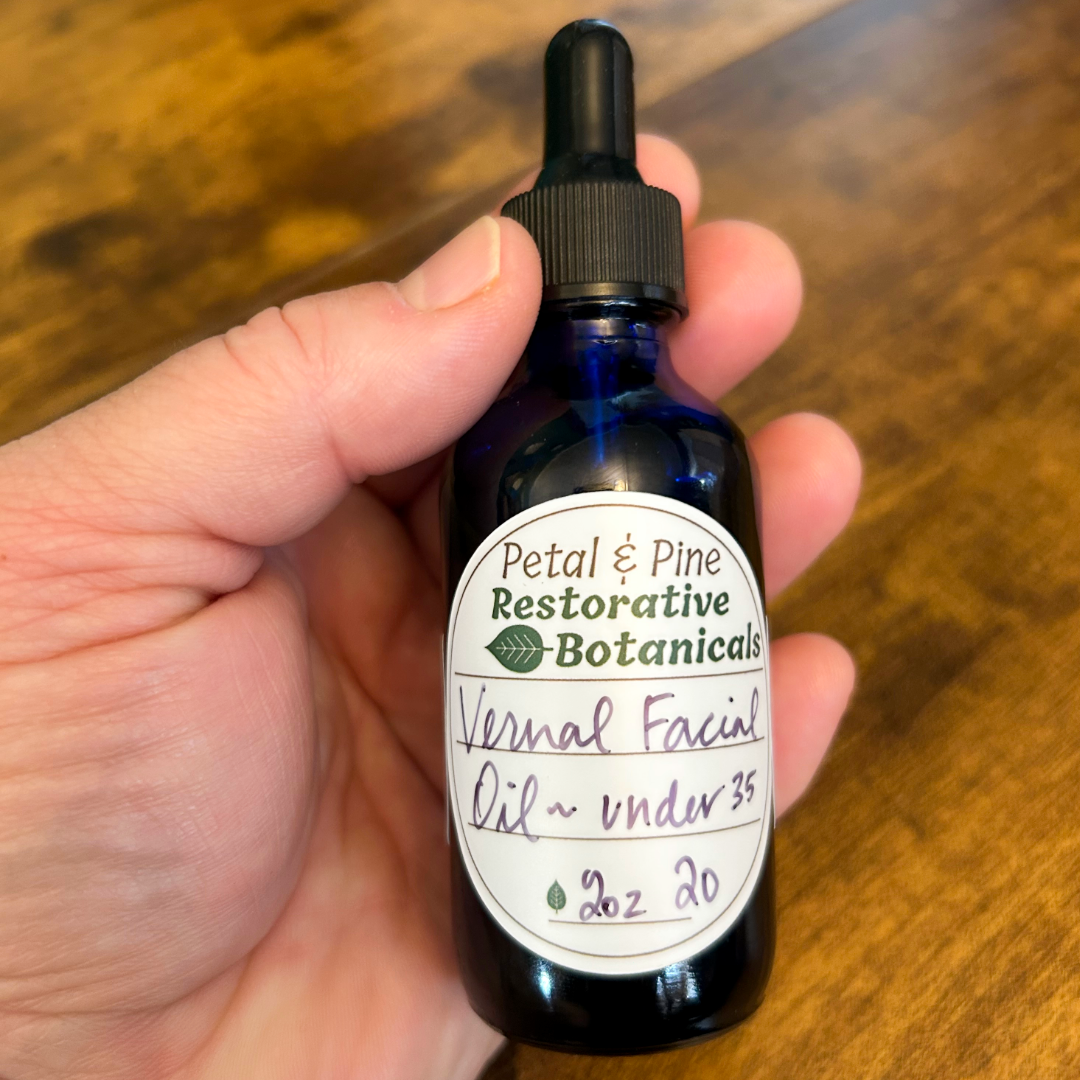 Hand holding a bottle of 'Petal & Pine Restorative Botanicals' Vernal Facial Oil on a wooden surface.