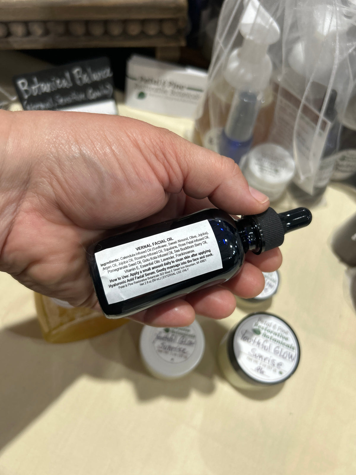 A hand holding a small black dropper bottle labeled 'Vernal Facial Oil' in a retail or workshop setting with other products blurred in the background.