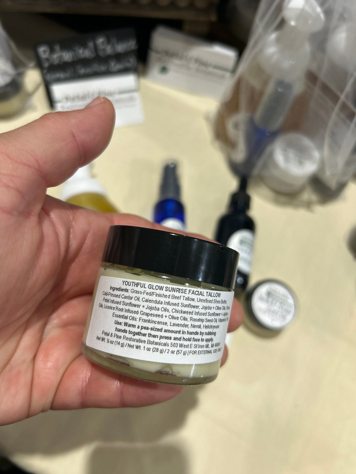A hand holding a small jar of Youthful Glow Sunrise Facial Salon treatment, showing the back of the label with ingredients and instructions, with a blurry salon counter and other bottles in the background.
