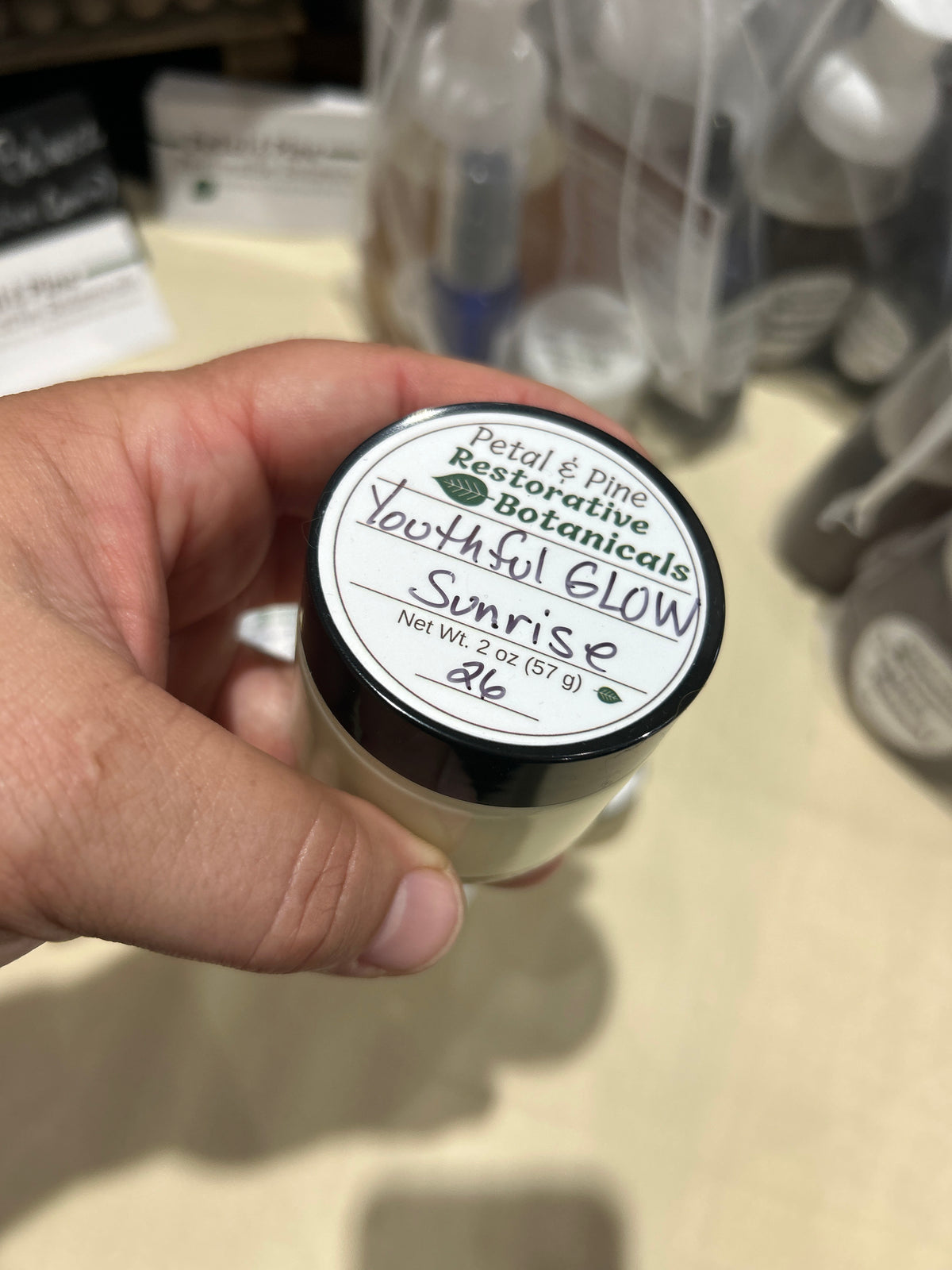 A hand holding a small jar of Petal & Pine Restorative Botanicals 'Youthful Glow' in the 'Sunrise' scent, with a blurry store shelf background.